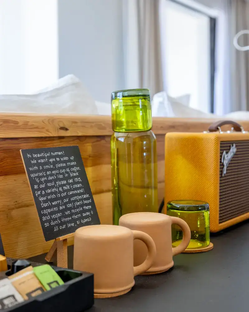 Wooden counter with two upside-down beige ceramic mugs, a green glass carafe with matching cups, a small chalkboard with handwritten message, and a yellow Fender speaker.