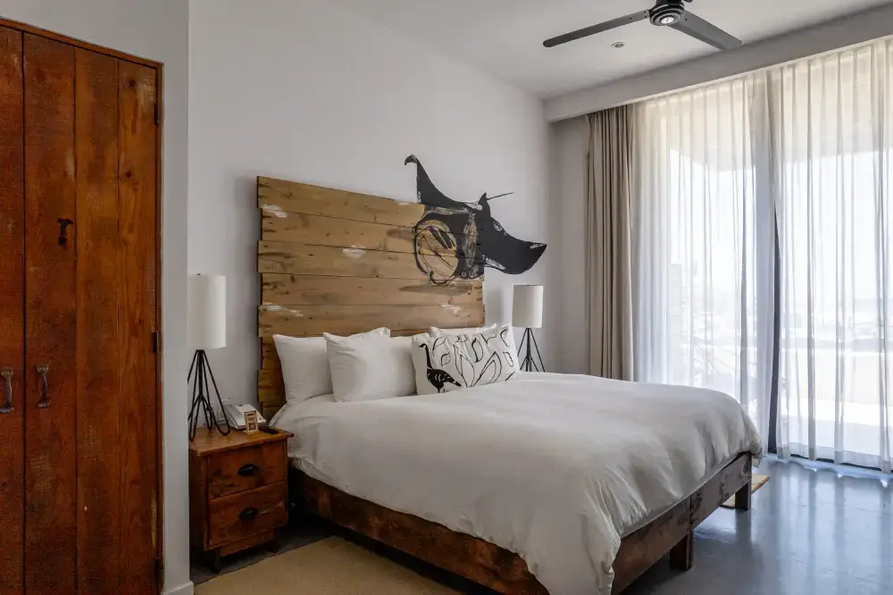 Minimalist bedroom with a wooden headboard featuring a black dog mural, white bedding, wooden nightstands with lamps, and sheer curtains letting in natural light.