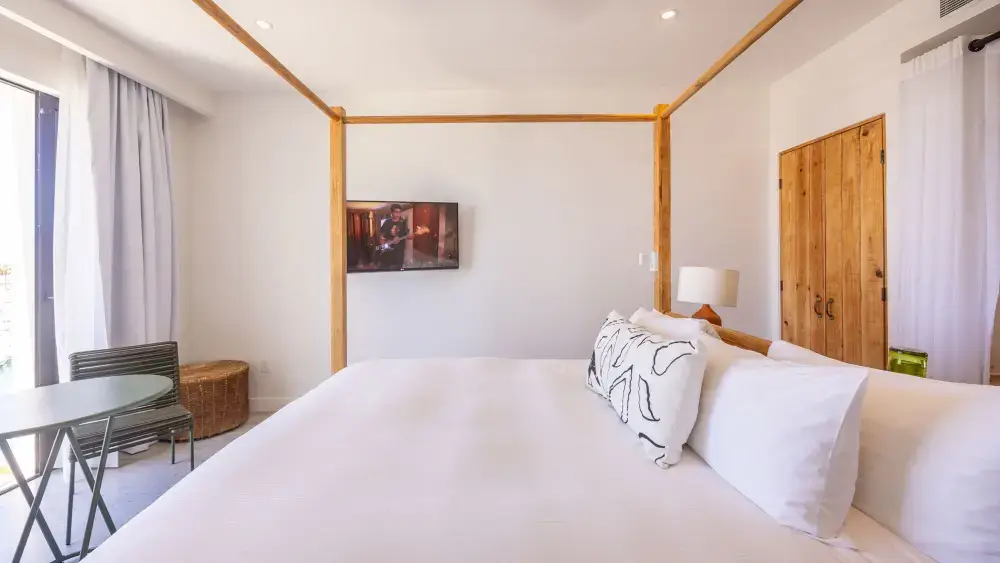 Bright bedroom with a four-poster wooden bed, white bedding, patterned pillows, a small TV mounted on the wall, a wicker basket, and a chair next to a window with white curtains.