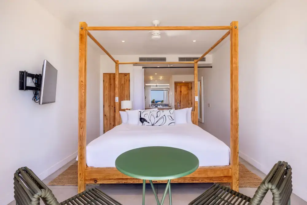 Bright bedroom with a wooden four-poster bed, white bedding, decorative pillows, a green round table, and two woven chairs, with wooden doors and a wall-mounted TV.