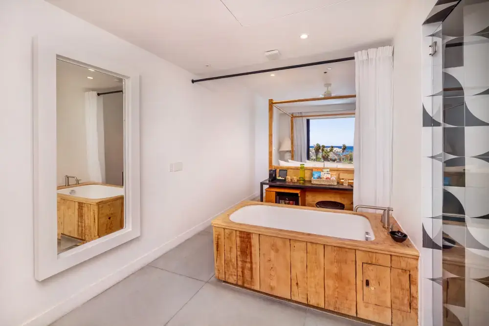 Minimalist bathroom with a wooden-framed bathtub, large mirror, and vanity table with ocean view through window.