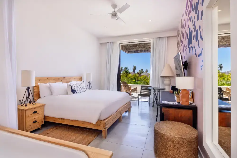 Habitación de hotel moderna y luminosa con una gran cama de madera, sábanas blancas, lámparas de noche, un balcón con sillas y vegetación tropical en el exterior.