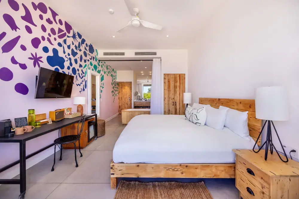 Dormitorio moderno con una gran cama de madera, sábanas blancas, lámparas de noche, un escritorio con una silla y un colorido mural abstracto en la pared.