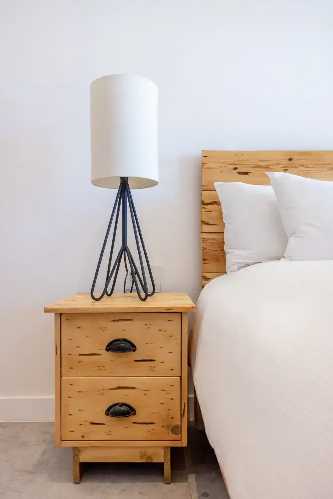 Mesita de noche de madera con dos cajones y una lámpara moderna con pantalla blanca junto a una cama con almohadas blancas y cabecero de madera.