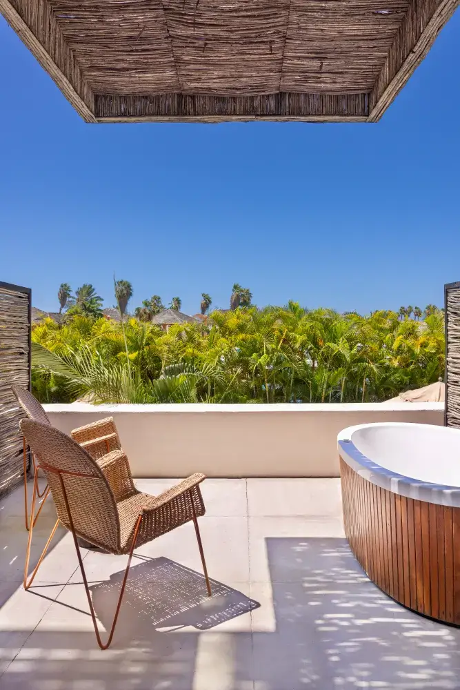 Patio exterior con dos sillas de mimbre y una bañera de hidromasaje de madera con vistas a exuberantes palmeras bajo un cielo azul claro.