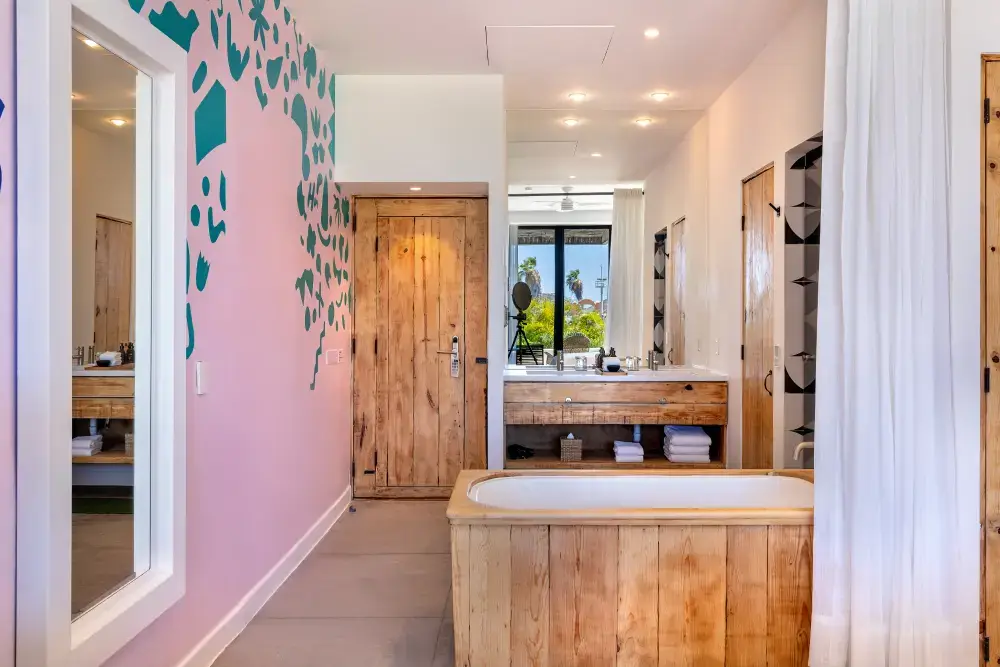 Cuarto de baño moderno con bañera de madera, espejo grande, puerta de madera, pared rosa con diseño abstracto verde azulado y cortina blanca.
