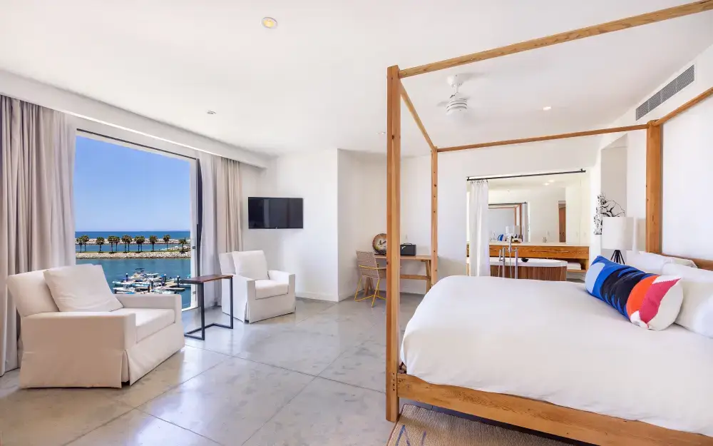 Bright hotel room with a large wooden canopy bed, white armchairs, a desk, and a window overlooking a marina and palm trees.