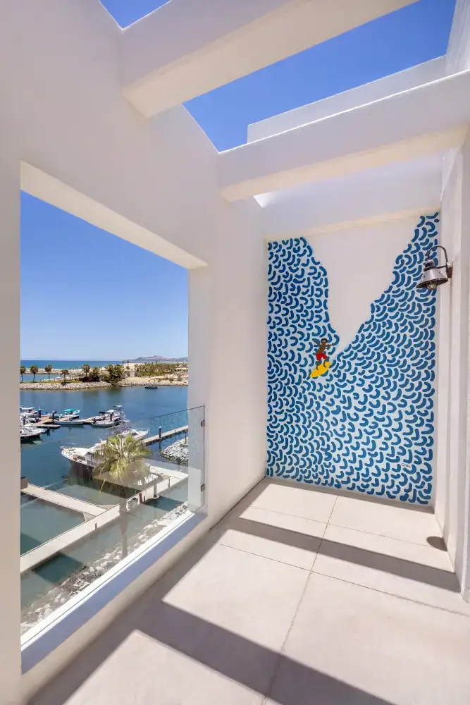 Balcony with white walls and decorative mural of a surfer on blue waves, overlooking a marina with boats and palm trees under a clear blue sky.