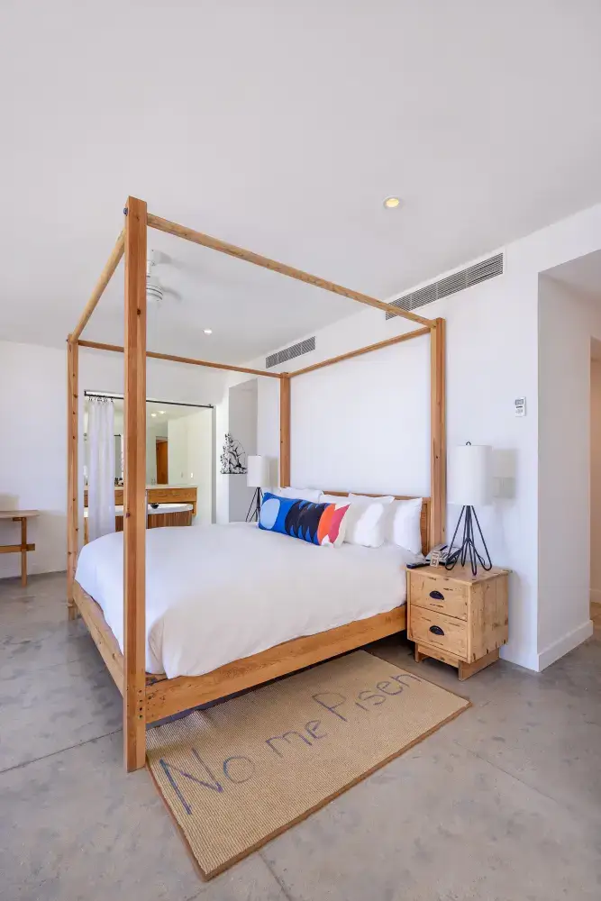 Bright bedroom with a wooden four-poster bed, white bedding, colorful pillows, two bedside lamps, and a rug with the text 'No me Piseh'.