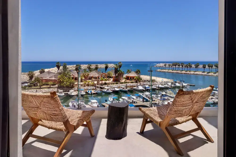 Two woven chairs and a small wooden table on a balcony overlooking a marina with boats, palm trees, and the ocean under a clear blue sky.