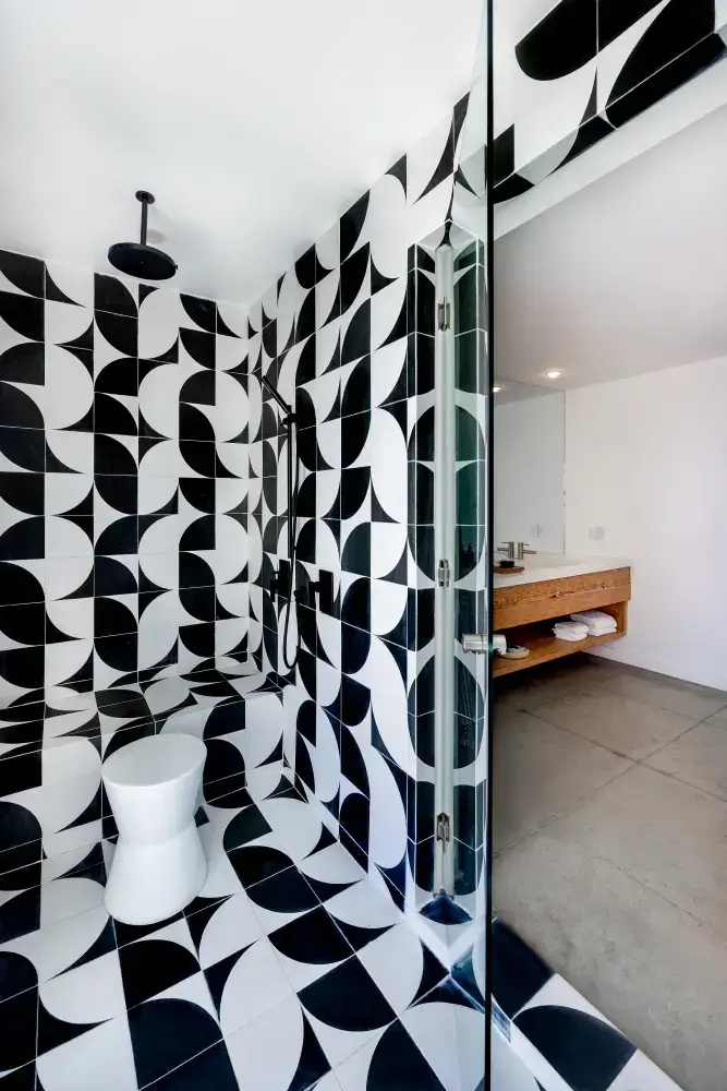 Modern shower area with black and white geometric patterned tiles, a built-in bench, and a white stool, adjacent to a bathroom with wooden vanity and concrete floor.