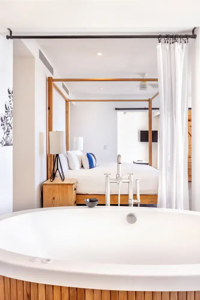 Modern bedroom with a wooden four-poster bed and white bedding seen through a large white bathtub in the foreground.