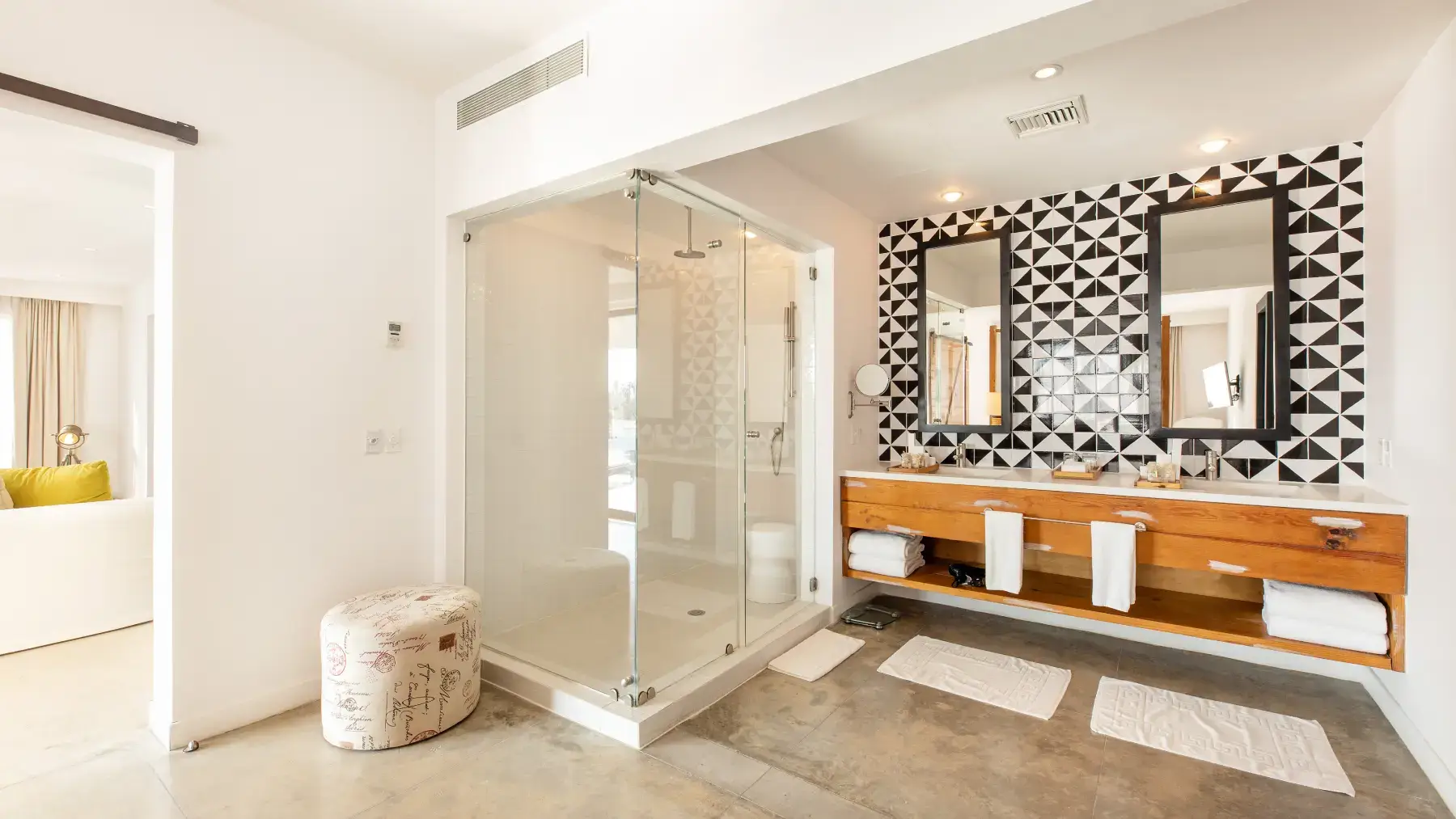 Cuarto de baño moderno con ducha acristalada, tocador doble de madera con dos espejos y pared con azulejos geométricos en blanco y negro.