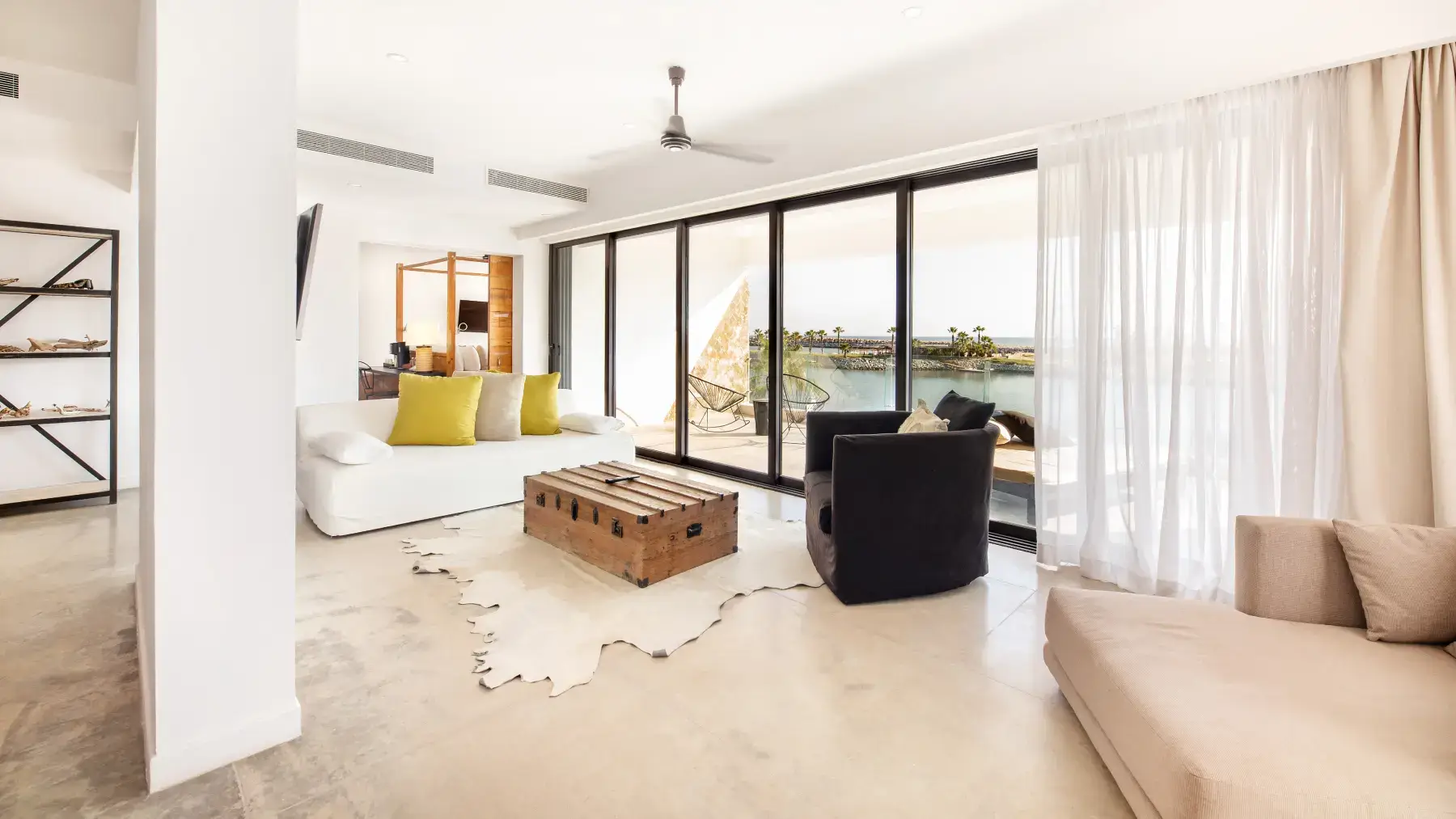 Luminoso salón con grandes puertas correderas de cristal con vistas al agua, con un sofá blanco con cojines verdes, un sillón negro, una mesa de centro de madera y una chaise longue beige.
