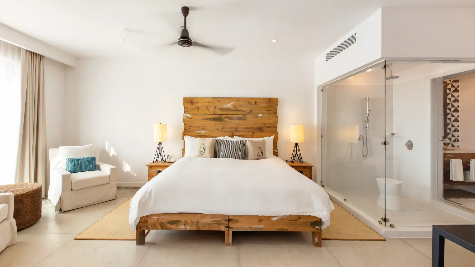 Dormitorio moderno con estructura de cama de madera, ropa de cama blanca, dos lámparas de mesilla, un sillón con cojín azul y una zona de ducha acristalada.