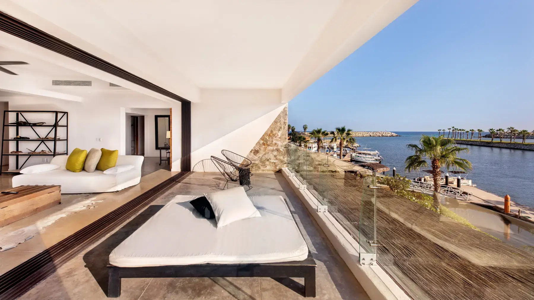 Moderna sala de estar abierta con sofá blanco y cojines amarillos que da a un balcón con diván y sillas con vistas a un puerto deportivo con palmeras y barcos bajo un cielo azul claro.