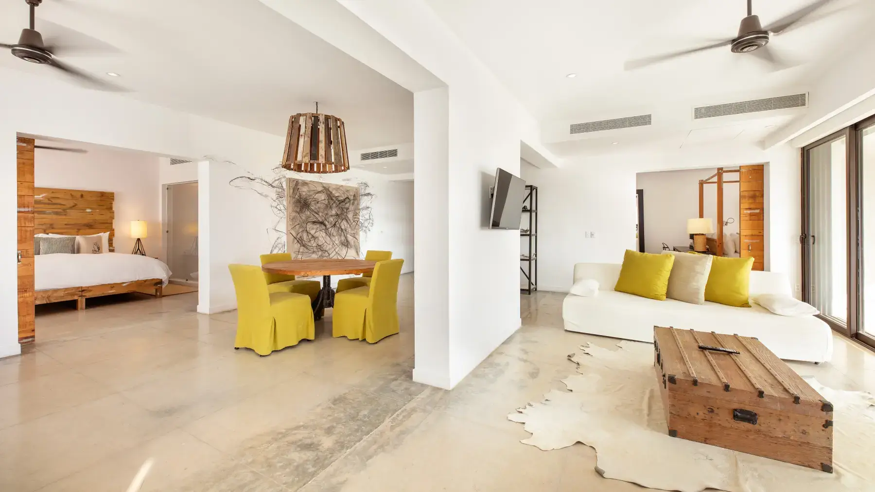 Moderno espacio abierto con paredes blancas, un sofá blanco con cojines de colores, mesa de centro de madera, mesa de comedor redonda con sillas amarillas y dos dormitorios con camas de madera.