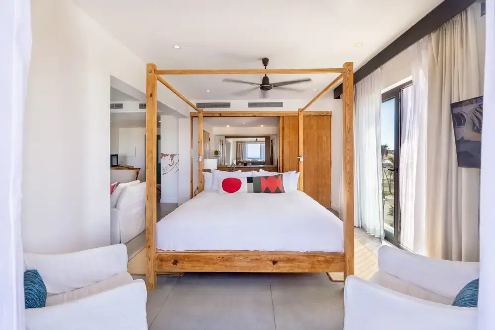 Habitación luminosa con cama de madera con dosel, ropa de cama blanca, cojines decorativos, dos sillones blancos y puertas correderas de cristal con cortinas transparentes.