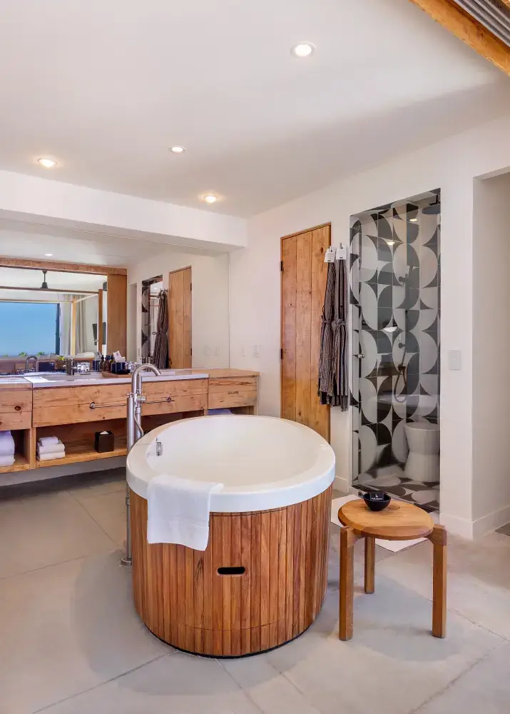 Cuarto de baño moderno con una bañera redonda de madera, un pequeño taburete de madera, un espejo grande y una mampara de ducha de cristal estampado.