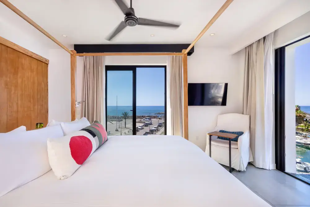 Habitación de hotel luminosa con una cama grande, almohadas de colores, ventilador de techo, televisor montado en la pared, sillón y vistas al mar a través de puertas correderas de cristal.