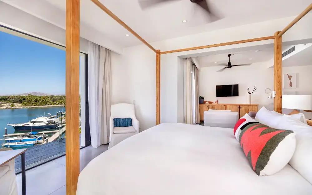Luminoso dormitorio principal con cama con dosel de madera, ropa de cama blanca y almohadas con estampado rojo, con vistas al puerto deportivo y a los barcos a través de puertas correderas de cristal.