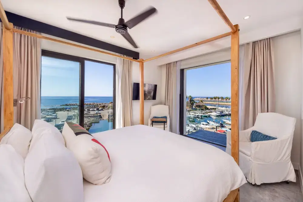 Habitación luminosa con cama con dosel de madera, sábanas blancas, dos sillones blancos y grandes ventanas con vistas al puerto deportivo y al océano.