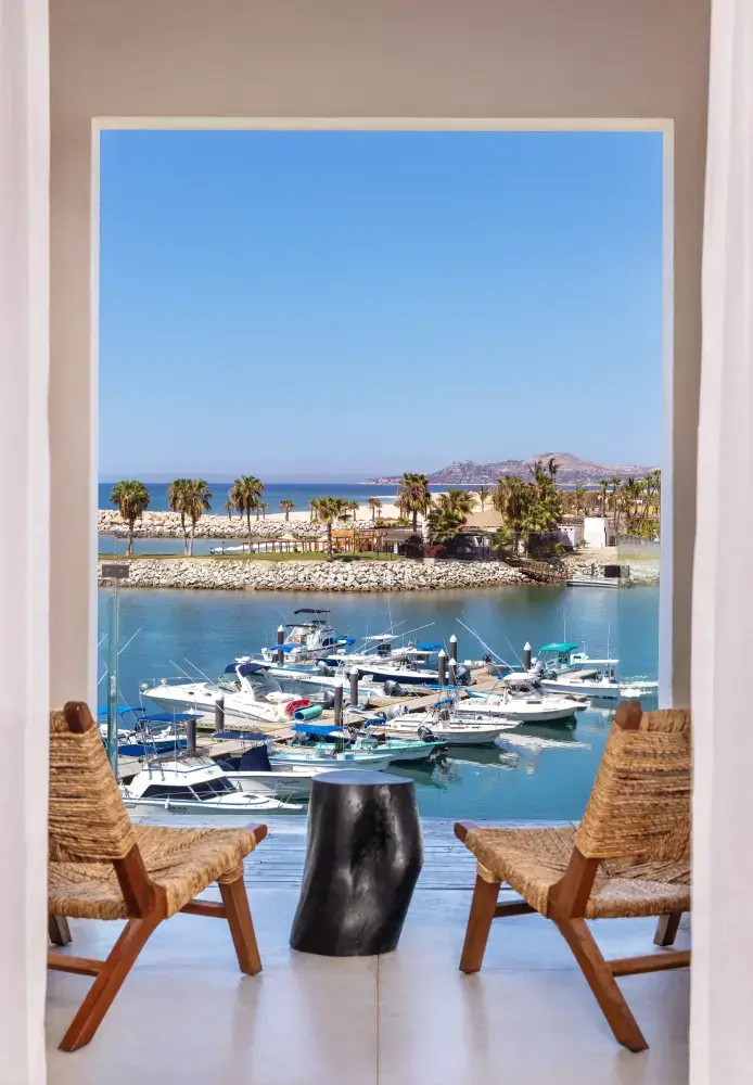 Vista a través de una puerta abierta que muestra dos sillas de mimbre y una pequeña mesa negra con vistas a un puerto deportivo con barcos y palmeras bajo un cielo azul claro.