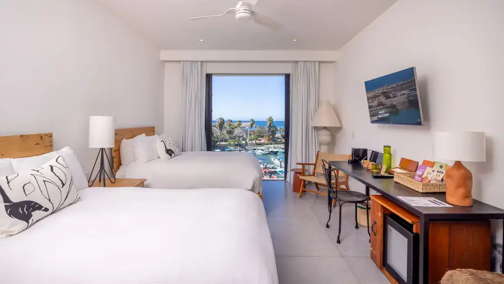 Habitación de hotel con dos camas blancas, un escritorio con aperitivos, lámparas y una gran ventana con vistas al puerto deportivo y al océano.