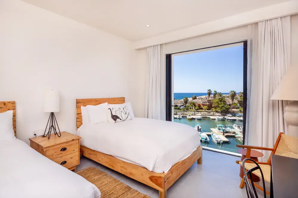Habitación de hotel luminosa con dos camas con estructura de madera, una mesita de noche con lámpara y una gran ventana con vistas al puerto deportivo con barcos y al océano.