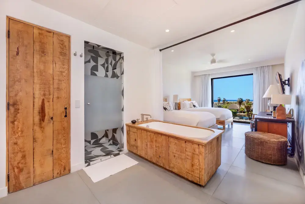 Habitación de hotel moderna con dos camas, bañera de madera, ducha con azulejos geométricos, puerta de madera y vistas al mar a través de un gran ventanal.