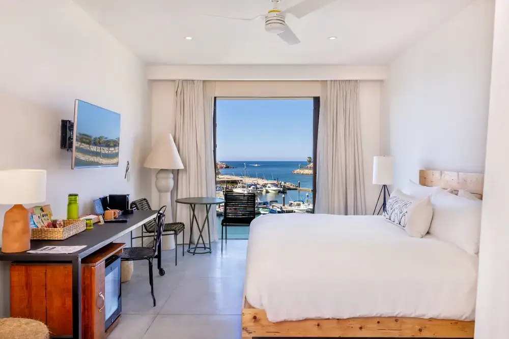 Bright hotel room with a large bed, desk area, and a window offering a view of a marina and ocean.