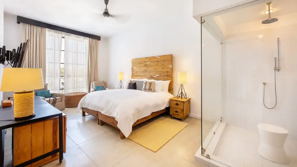 Bright modern bedroom with a wooden bed frame, white bedding, two bedside lamps, armchairs by a large window, and an open glass shower area.