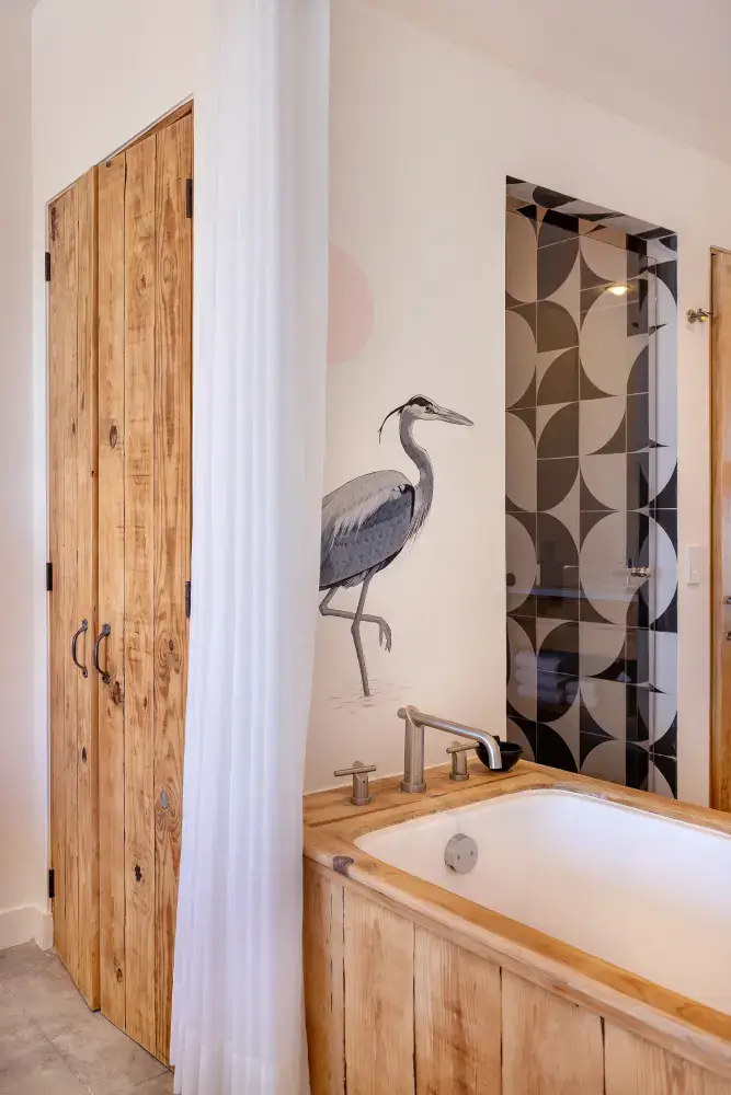 Bathroom with wooden-framed bathtub, wood-paneled closet doors, a shower with black and gray geometric tiles, and a heron mural on the wall.