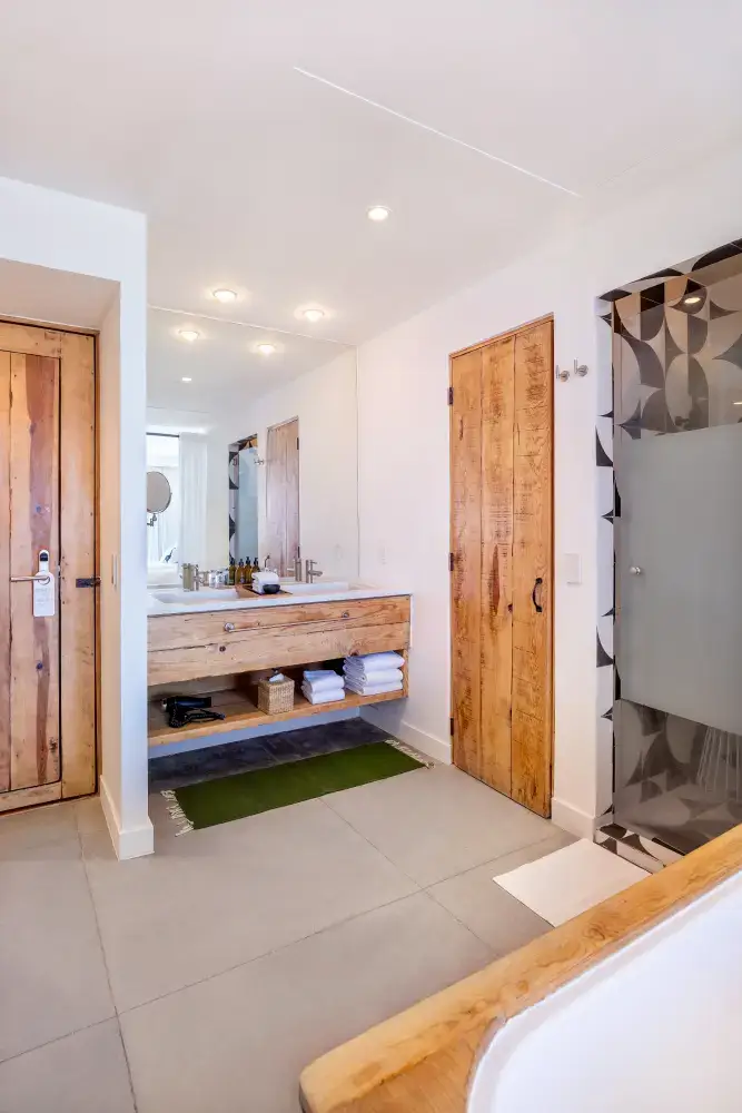 Cuarto de baño moderno con puertas de madera, un tocador flotante de madera con dos lavabos y toallas debajo, un espejo grande, una puerta de ducha de cristal esmerilado con azulejos estampados y alfombrillas verdes y blancas.