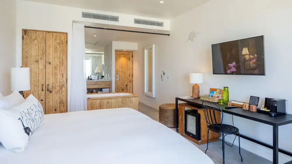 Modern hotel room with white bed, wooden furniture, a desk with chair, wall-mounted TV, and a bathtub near the entrance.