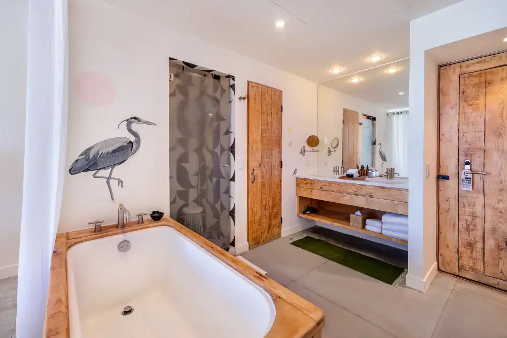 Cuarto de baño moderno con bañera con marco de madera, mural de una garza en la pared, puerta de ducha de cristal, lavabo de madera con toallas y espejo grande.