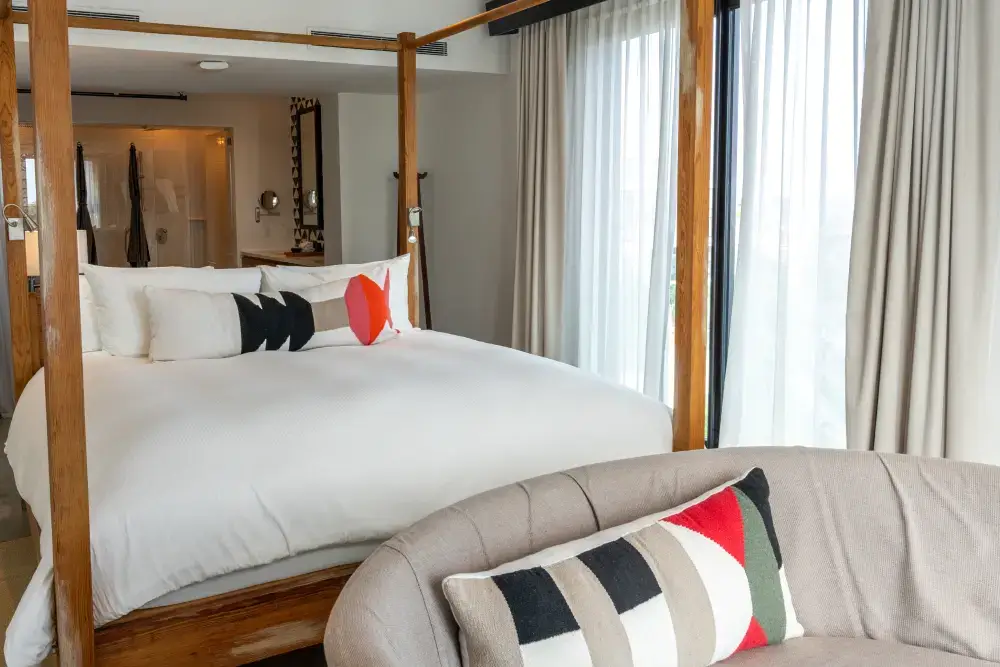 Modern hotel room with a wooden four-poster bed, white linens, decorative pillows, and a beige curved sofa near large windows with sheer curtains.
