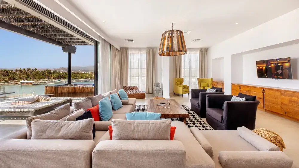 Spacious modern living room with beige sectional sofa, blue and red pillows, black armchairs, wooden coffee table, large windows, and a view of palm trees and water outside.