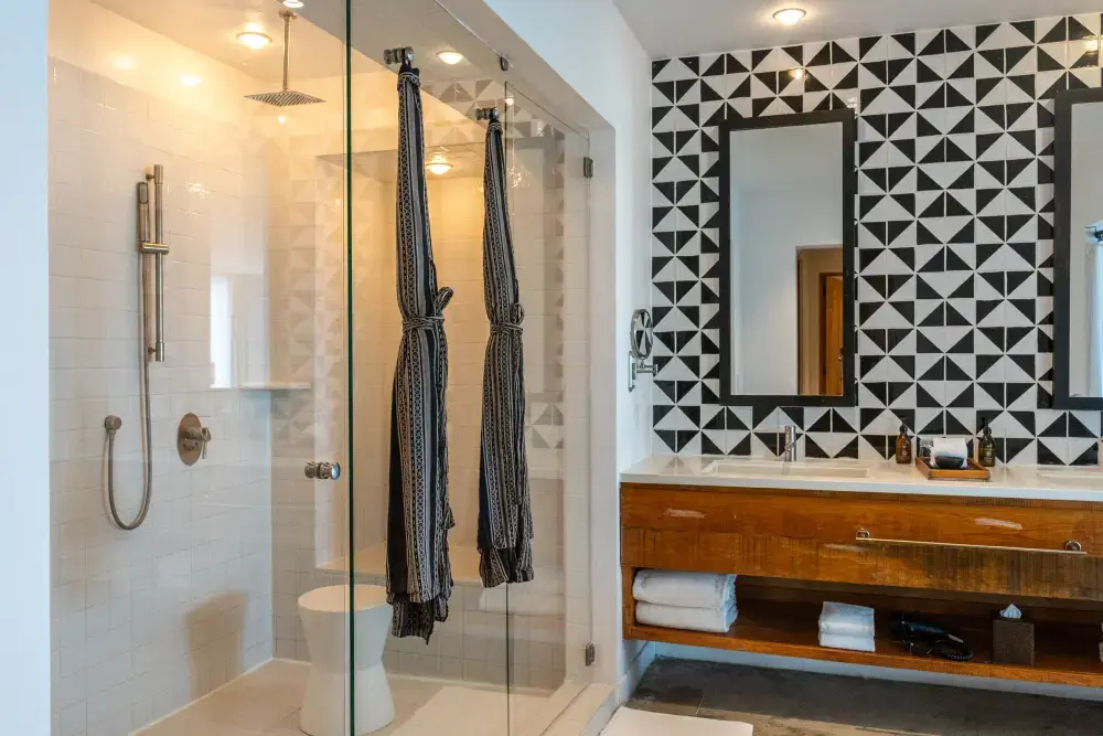 Modern bathroom with glass shower enclosure, dual mirrors, wooden vanity with towels, and black and white geometric tile wall.