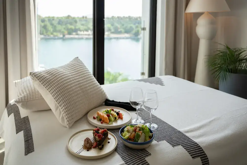 Bed with textured white pillows, plates of fruit, salad, dessert, two empty wine glasses, and a large window showing a lake view in the background.