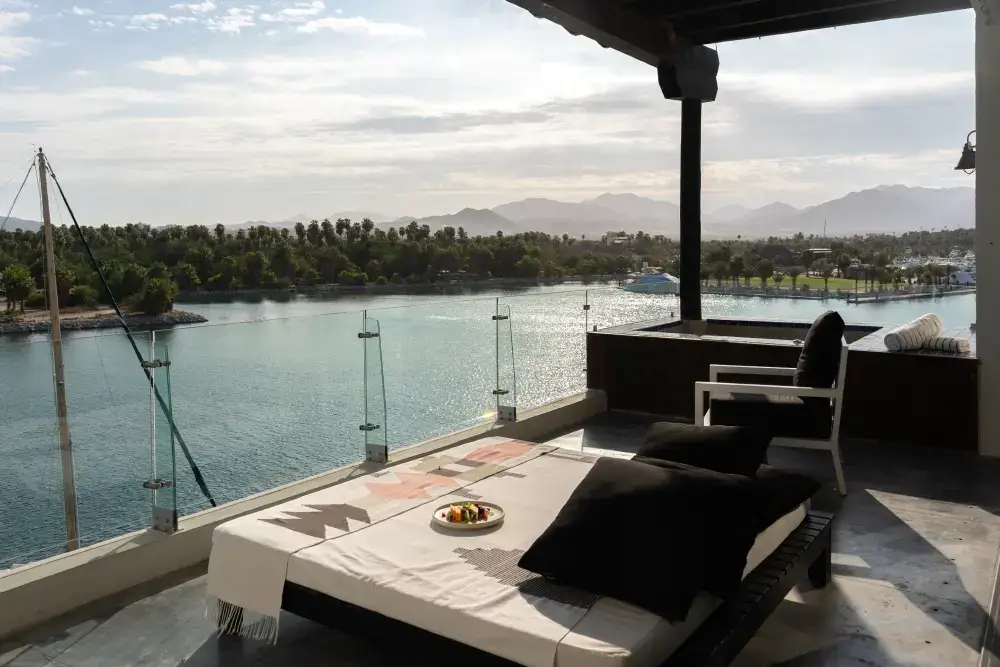 Balcony with a daybed and chair overlooking a calm water body, palm trees, boats, and mountains in the distance under a partly cloudy sky.