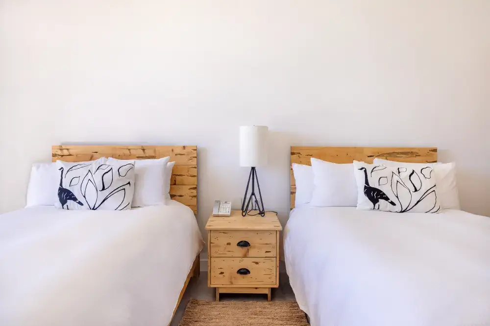 Dos camas perfectamente hechas con sábanas blancas y almohadas decorativas con diseños de pájaros negros, separadas por una mesita de noche de madera con una lámpara moderna y un teléfono.