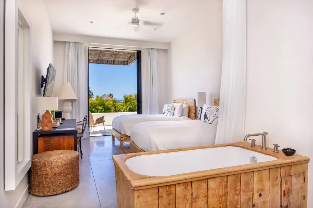 Habitación de hotel luminosa con dos camas blancas, una bañera de madera, un escritorio con lámparas y puertas de patio abiertas que dan a una zona verde exterior.