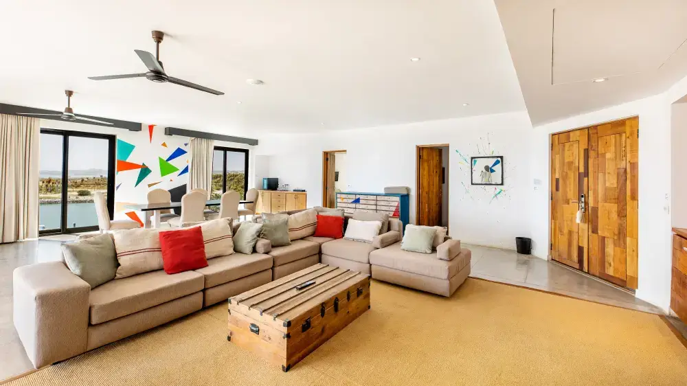 Luminoso salón con un gran sofá modular beige, mesa de centro de madera, mesa de comedor con sillas y coloridos cuadros geométricos cerca de las ventanas con vistas al agua.