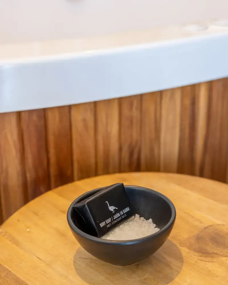 Pequeño cuenco negro con cristales gruesos de sal de baño blanca y una pastilla de jabón corporal envuelta en papel negro sobre una superficie de madera.