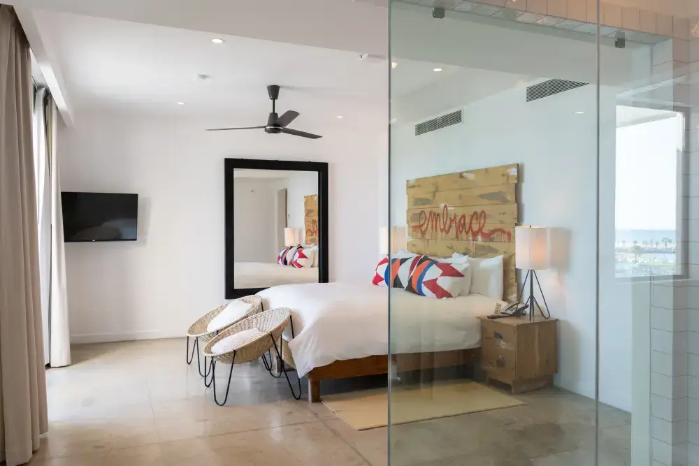 Dormitorio moderno con cabecero de madera con la palabra «embrace», ropa de cama blanca con almohadas de colores, dos sillas de mimbre, una lámpara de pie, un gran espejo de pared y un televisor montado en la pared.