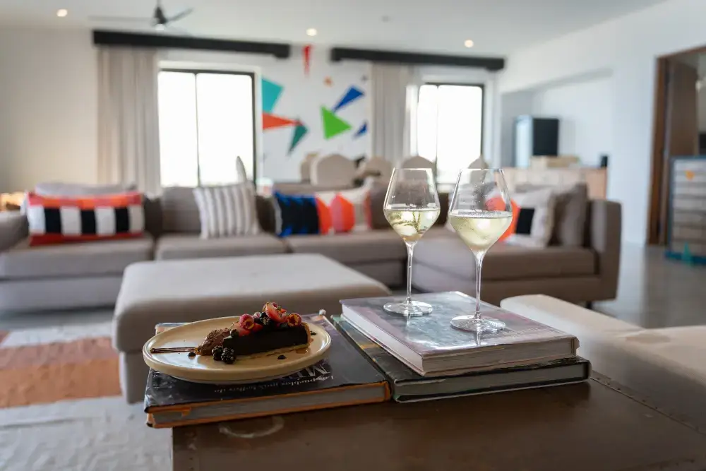Dos copas de vino blanco y un plato con un postre de chocolate cubierto con bayas sobre unos libros en una sala de estar moderna.