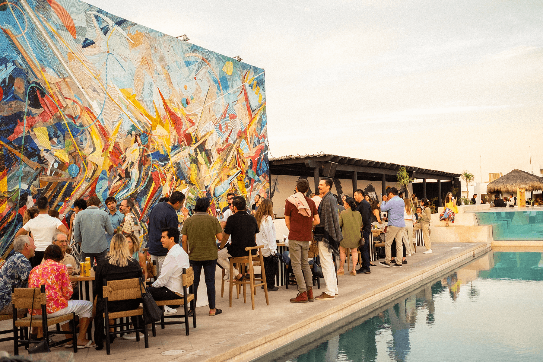 People socializing at an outdoor poolside event with a colorful abstract mural on the wall.