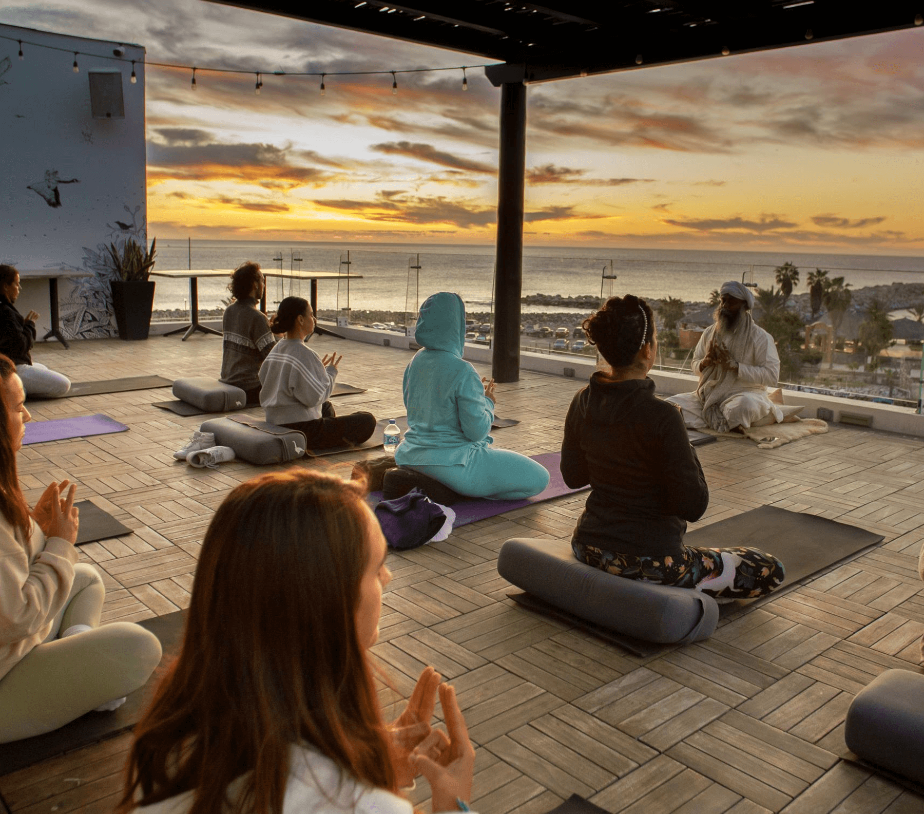 Group meditation session at sunset on a wooden deck overlooking the ocean, with participants sitting on cushions facing a seated instructor.
