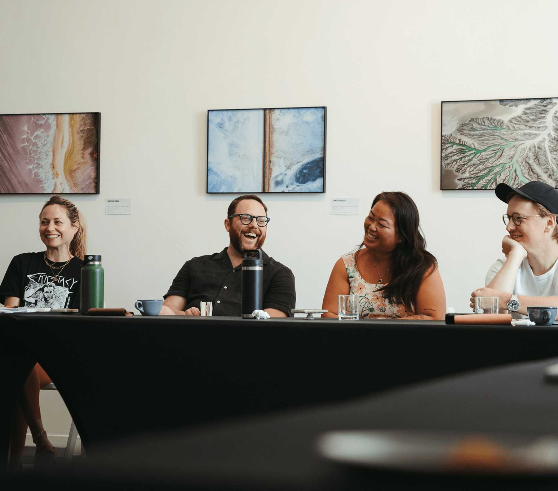 Four people sitting at a table in a gallery space, smiling and laughing with abstract landscape artwork on the wall behind them.